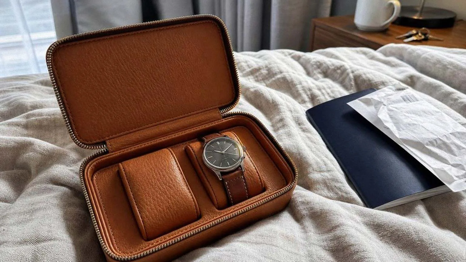 Estuche de viaje de piel marrón con cremallera abierto sobre una cama, con dos huecos acolchados y un reloj de correa marrón